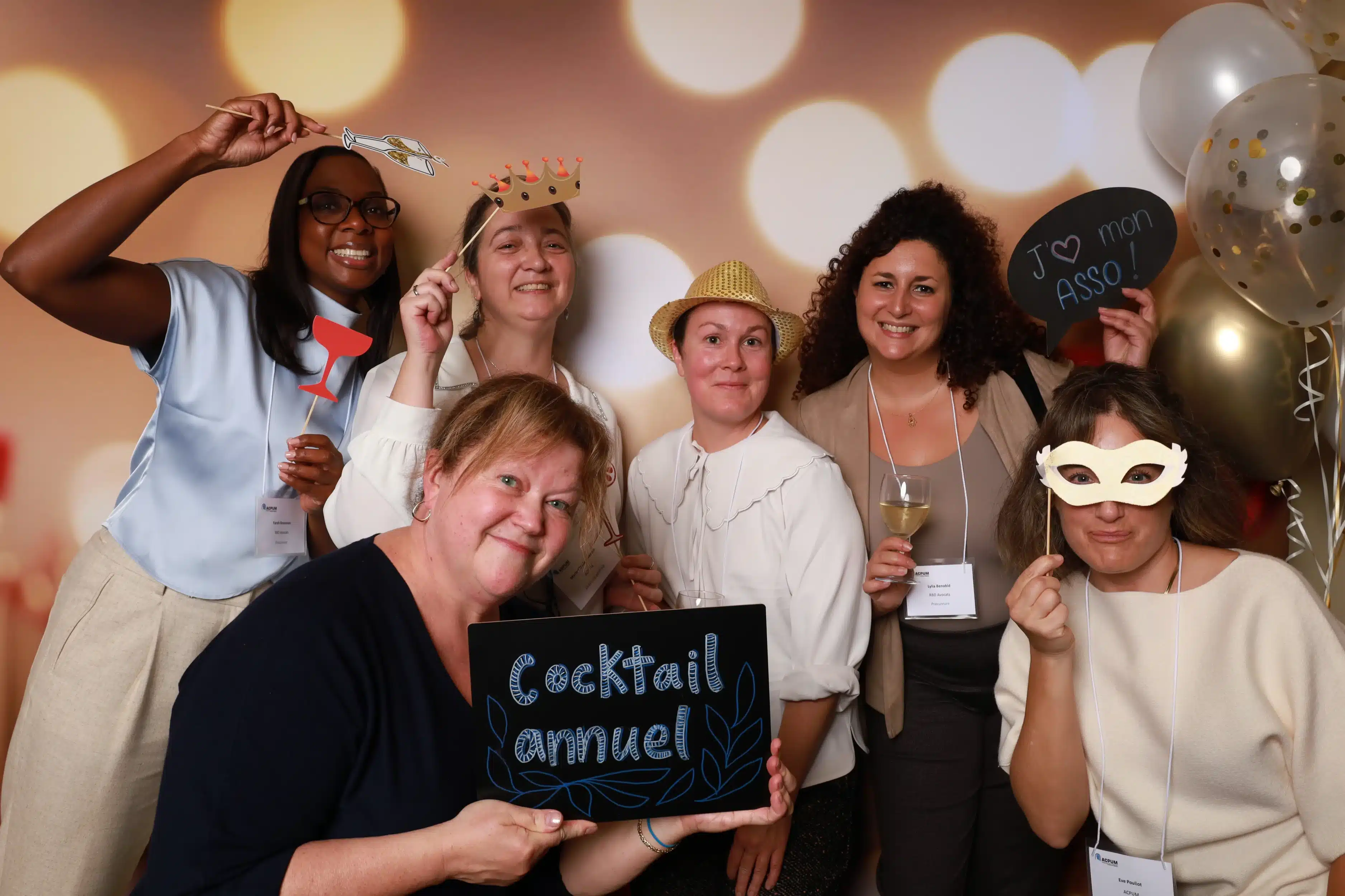 Groupe de six personnes dans un photobooth CheeeseBOX, souriantes avec divers accessoires comme des lunettes, chapeaux et panneaux, sur fond festif.