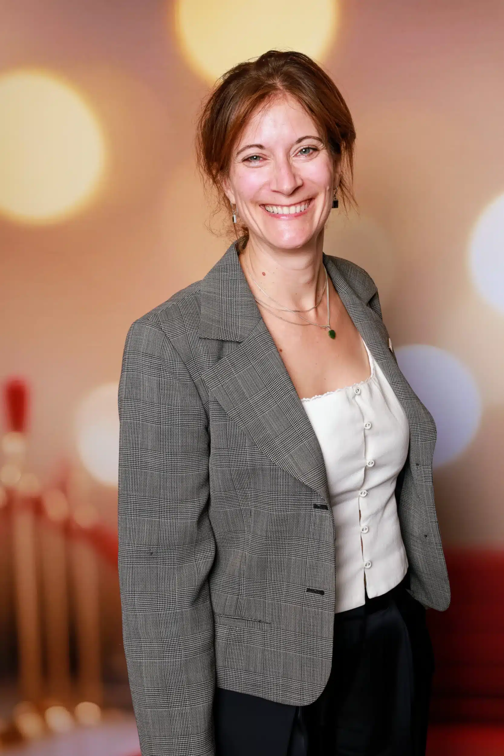 Femme souriante dans un photobooth CheeeseBOX, portant un blazer et un top, avec un fond flou aux lumières colorées.